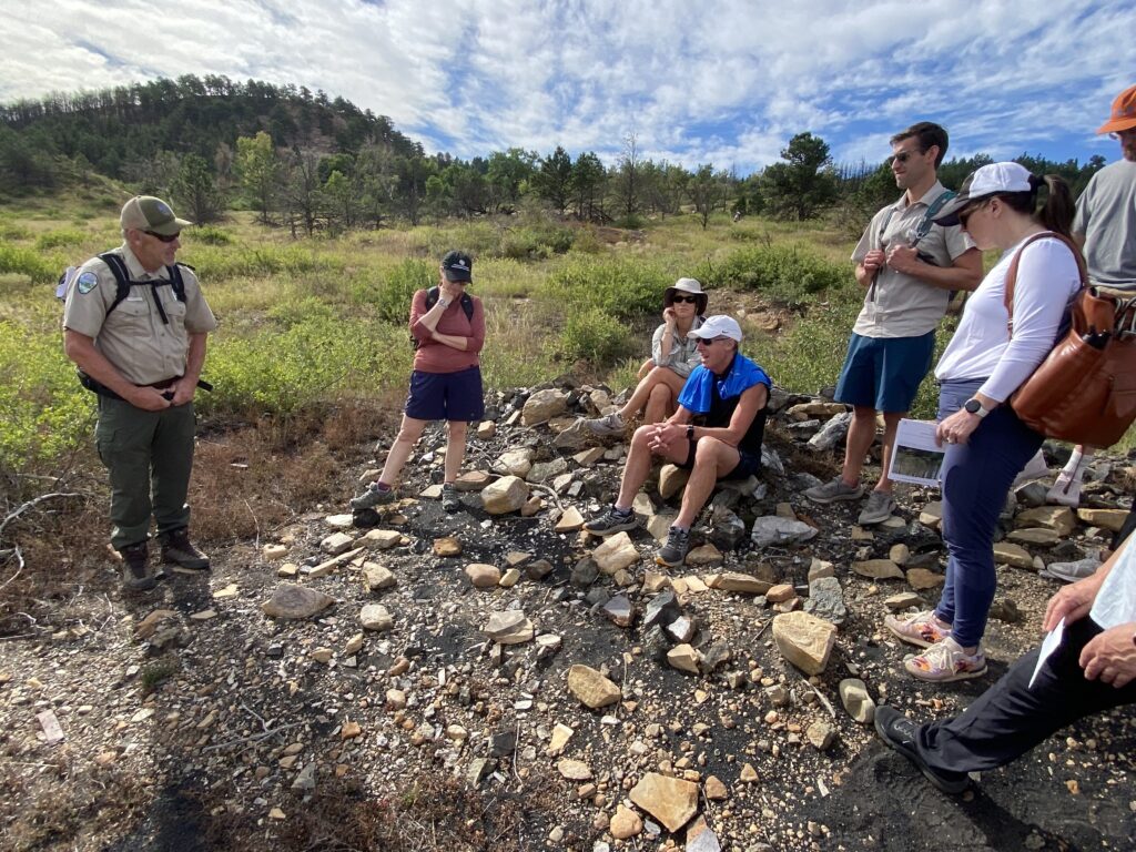 Students, standing in open space greenery and others sitting on rocks listen to a park ranger.