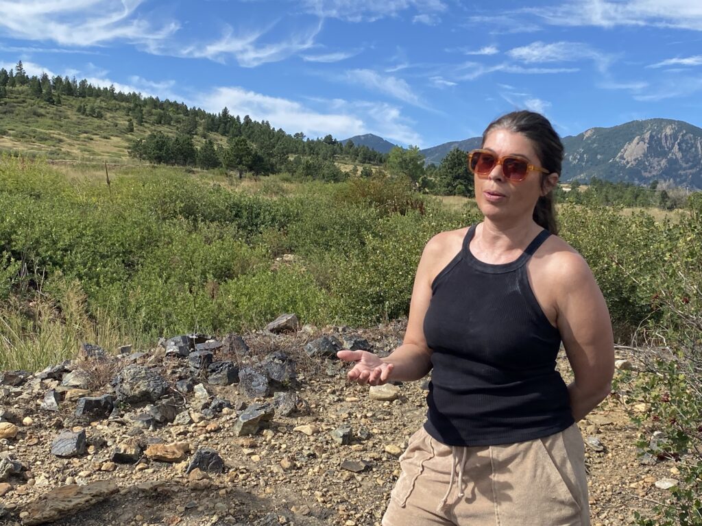 A woman in a black tank top speaks in the foreground and the Flatirons and Boulder's rolling green open space in the background.