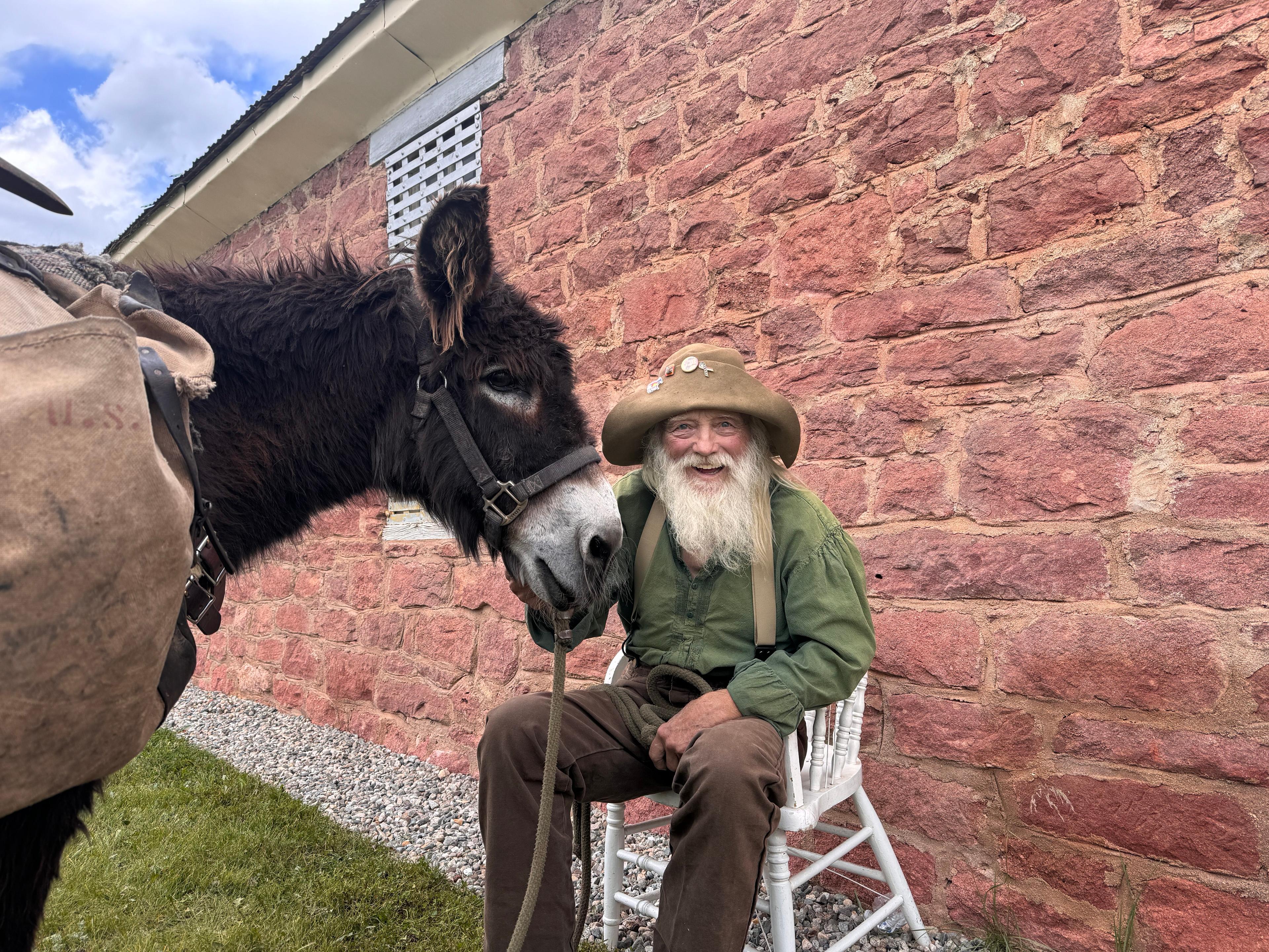 An old man in suspenders, a green shirt, and a brimmed hat sits against a brick wall. He holds the bridle of his large brown donkey.