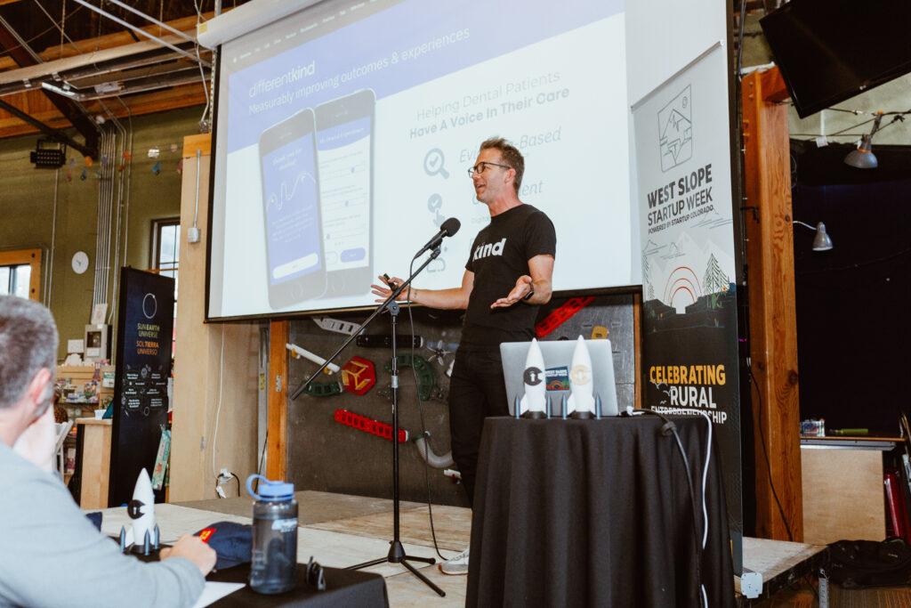 A man in a black shirt stands at a microphone stand holding out his open hands in a gesture. Behind him is a projection screen and a banner for West Slope Startup Week.