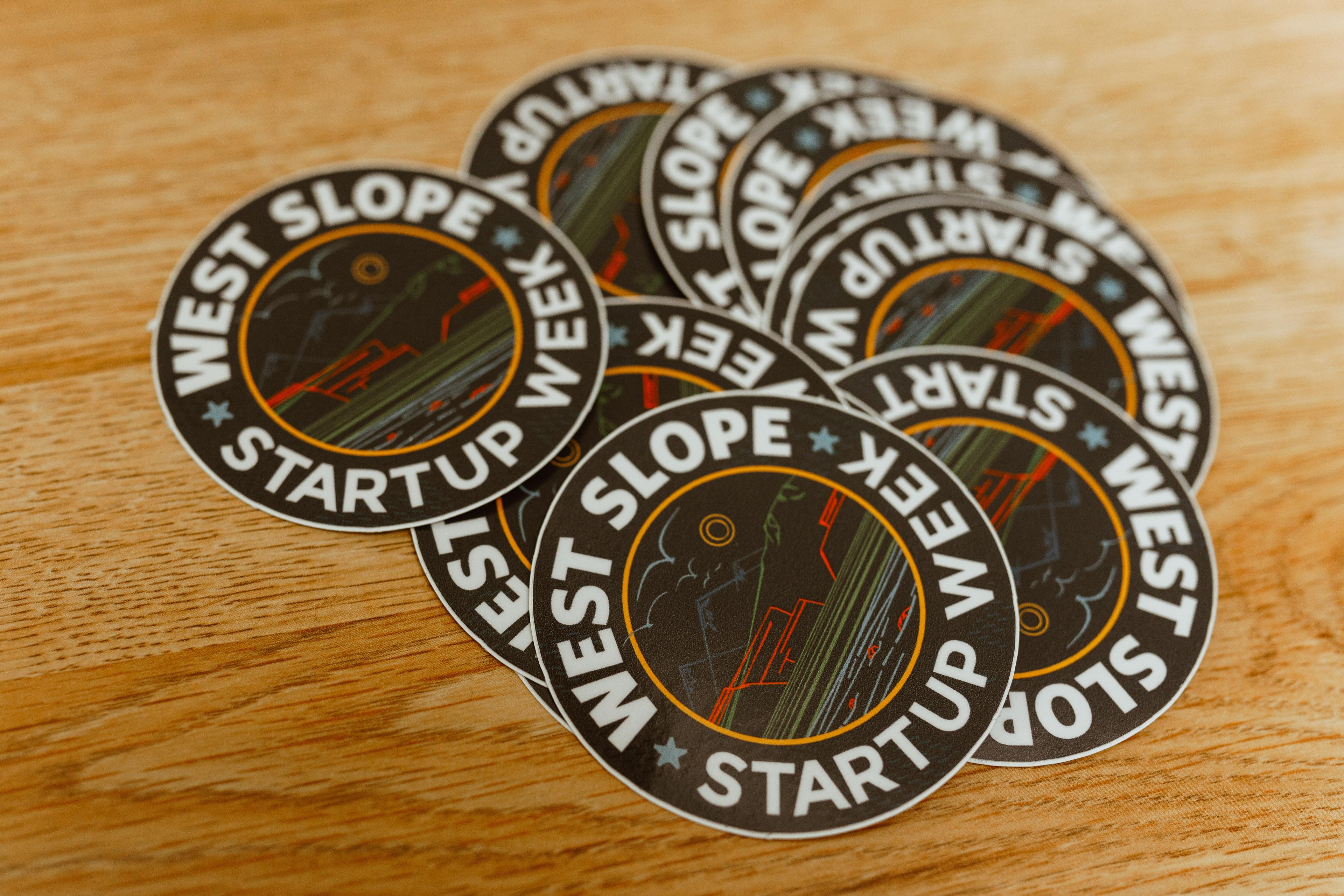 A pile of round stickers that say "West Slope Startup Week" sit on a table.