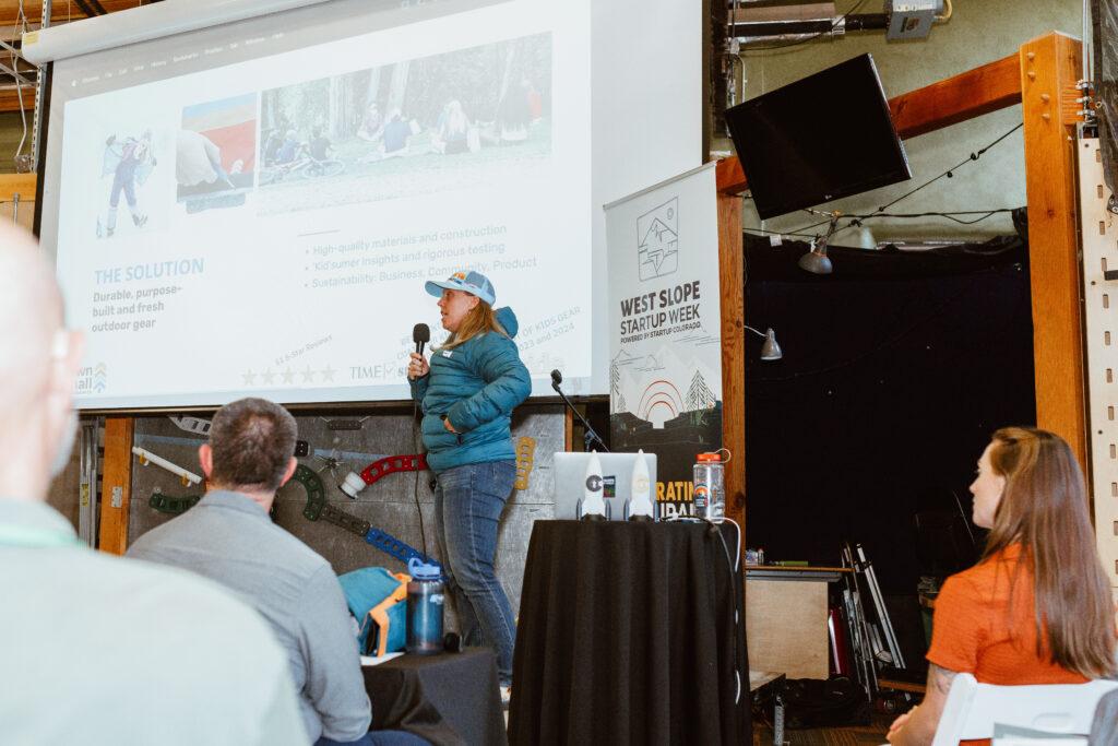 A woman in a blue cap and a blue puffy jacket holds a microphon with one hand and has her other hand in her pocket. Behind her is a projection screen and a banner for West Slope Startup Week. People are sitting down listening in the foreground.