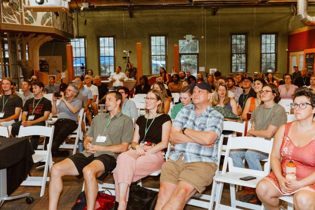 A crowded room of people sitting in rows attending the West Slope Startup Week. Everyone is looking forward, wearing green event lanyards.