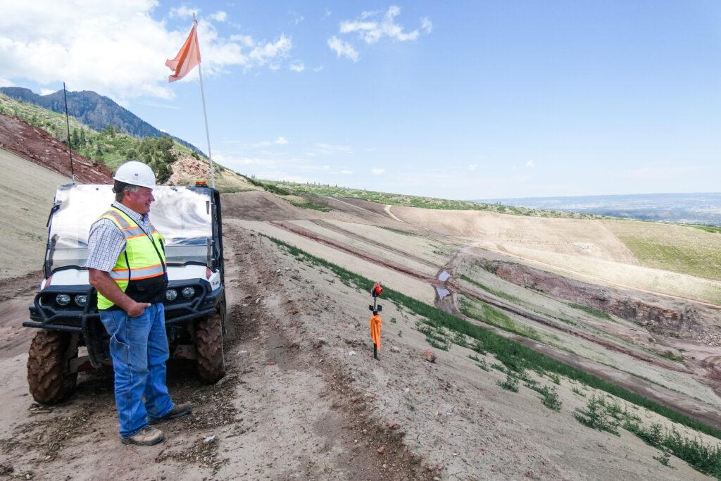 A man wearing a construction hardhat and safety vest stands in front of an open ATV that is parked at the top of a steep slope.