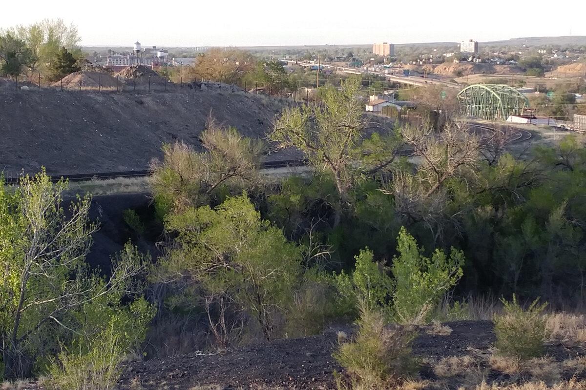 Trees and shrubs with green leaves grow in scattered spots from an area covered in black and dark gray rocks. Railroad tracks run behind it and beyond that is a steep embankment, I25, a bridge with arched light green girders a portion of the Pueblo cityscape.