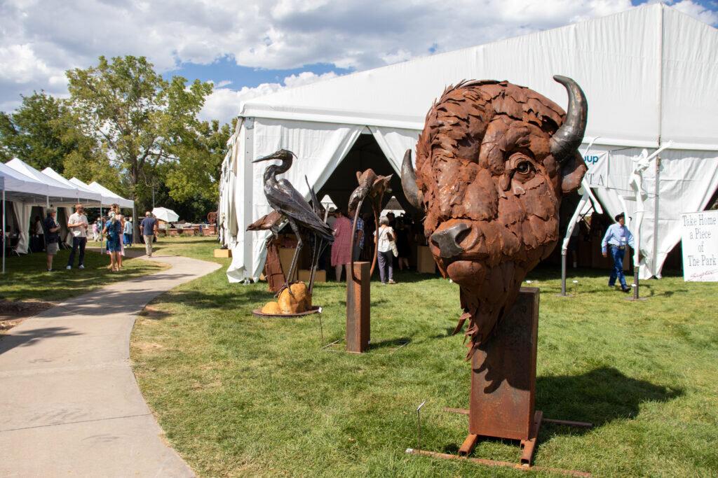 Works, including a large metal bison head and a Great Blue Heron, on display outside an event tent at the 2023 Loveland Sculpture in the Park festival.