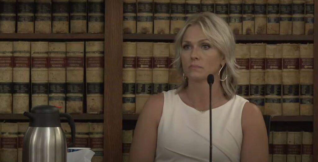 A blonde woman named Sherronna Bishop in a white dress sits at the witness stand in a courthouse in Grand Junction with a bookshelf containing law books in the background. A stainless steel pitcher of water sits on the table to Bishop's right.