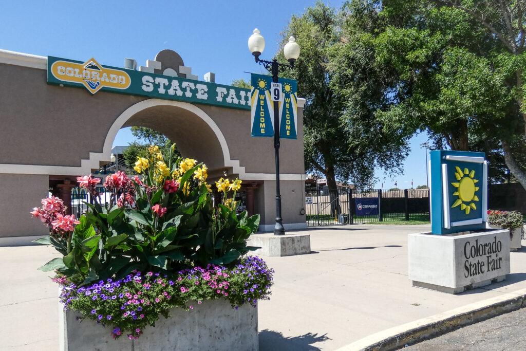 A green sign with white lettering says Colorado State Fair in western style font and a yellow background for the world Colorado is at the top of a neutral toned stucco structure featuring a large arched entry way with light colored trim. Another sign with a yellow sunburst on a green background stands near the road atop a concrete box with the words Colorado State Fair incised on it. Concrete boxes are filled with colorful flowers. Welcome banners hang from an antique style lamp post and trees grow near the gate.