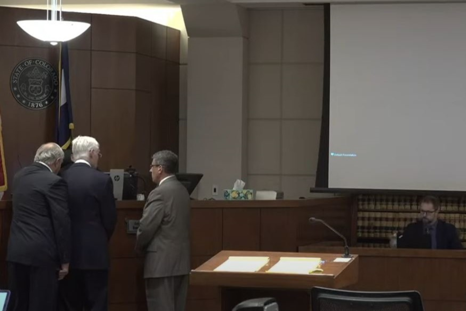 Three attorneys in suits gather at the judge's bench in a courtroom in Mesa County while a witness in white waits for questioning at the witness stand.