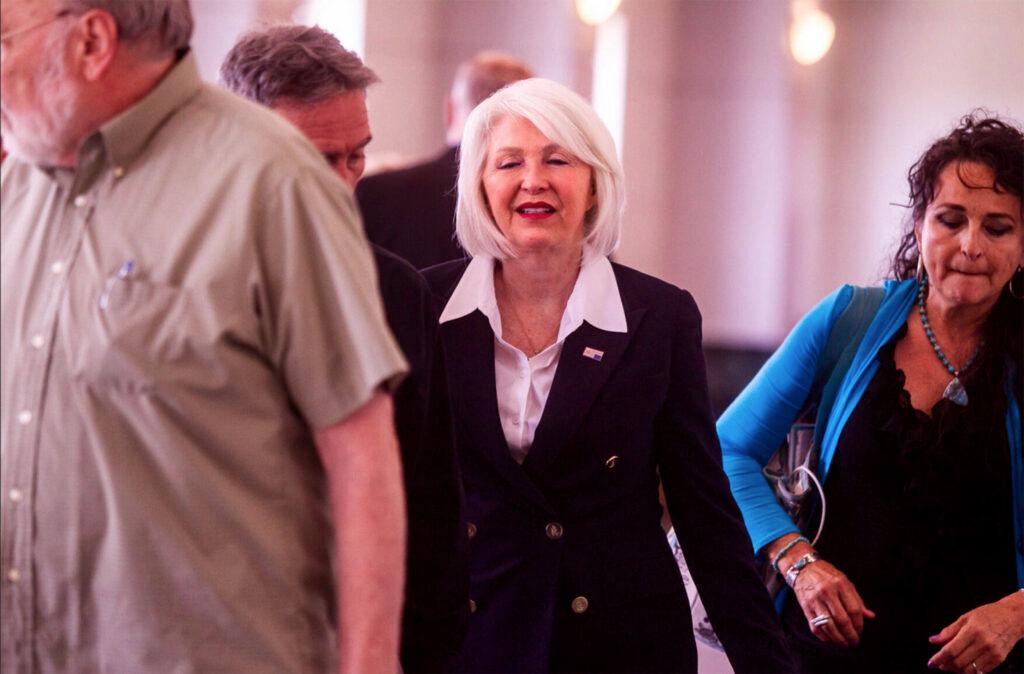 Tina Peters, wearing a blue double-breasted blazer and white shirt, walks down a white hallway outside a courtroom, flanked by a supporter wearing a blue shirt.