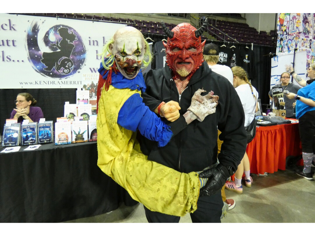 A man in a demon mask holding an animatronic killer clown on his side.