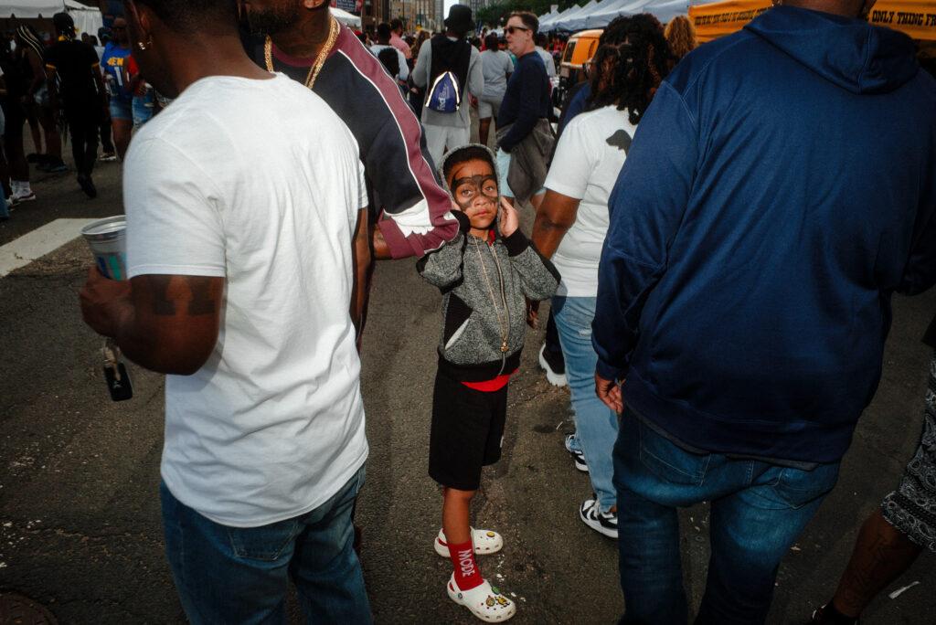 A candid photograph of a Black boy with Batman face paint, looking at the photographer through a crowd.