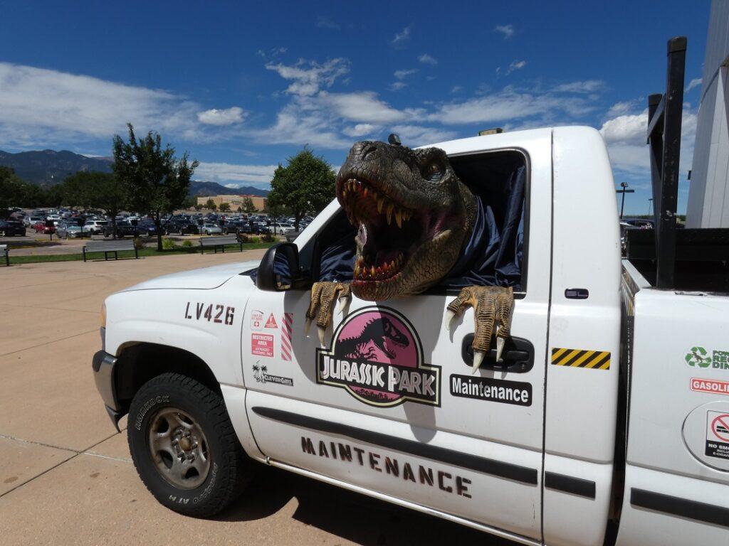 A fake dinosaur head sticks out of the driver side window of a truck