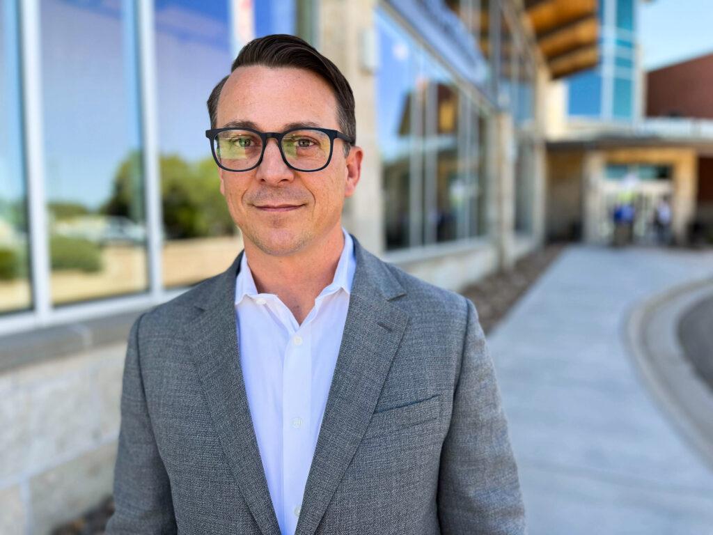 A man in a gray jacket and black glasses who running for a board seat poses for a photo near a building.