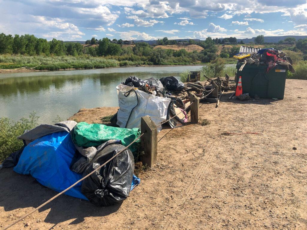 Bags and piles of trash line a river bank under fluffy clouds.