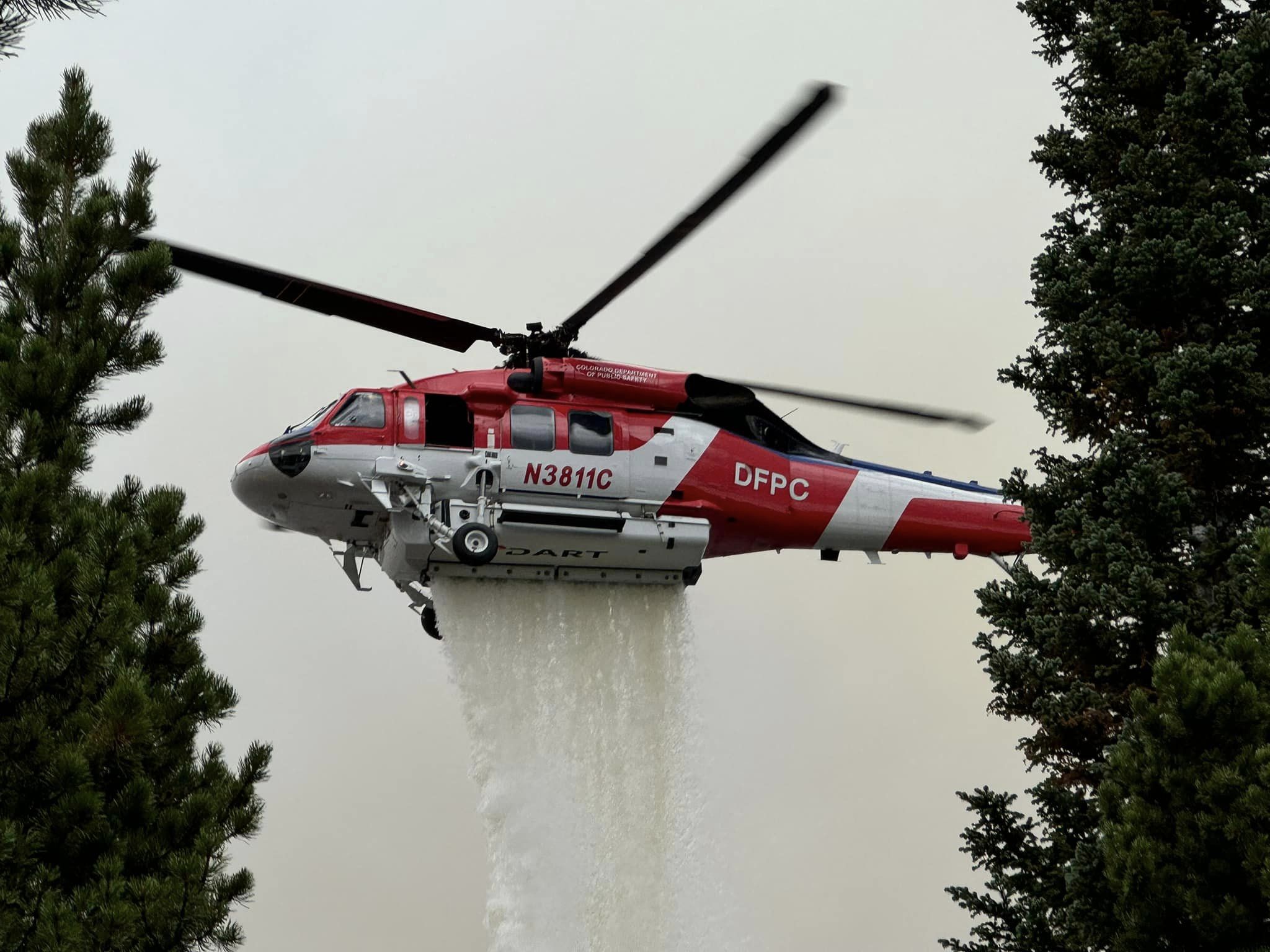 A red and white helicopter framed by two evergreen trees drops water on a wildfire