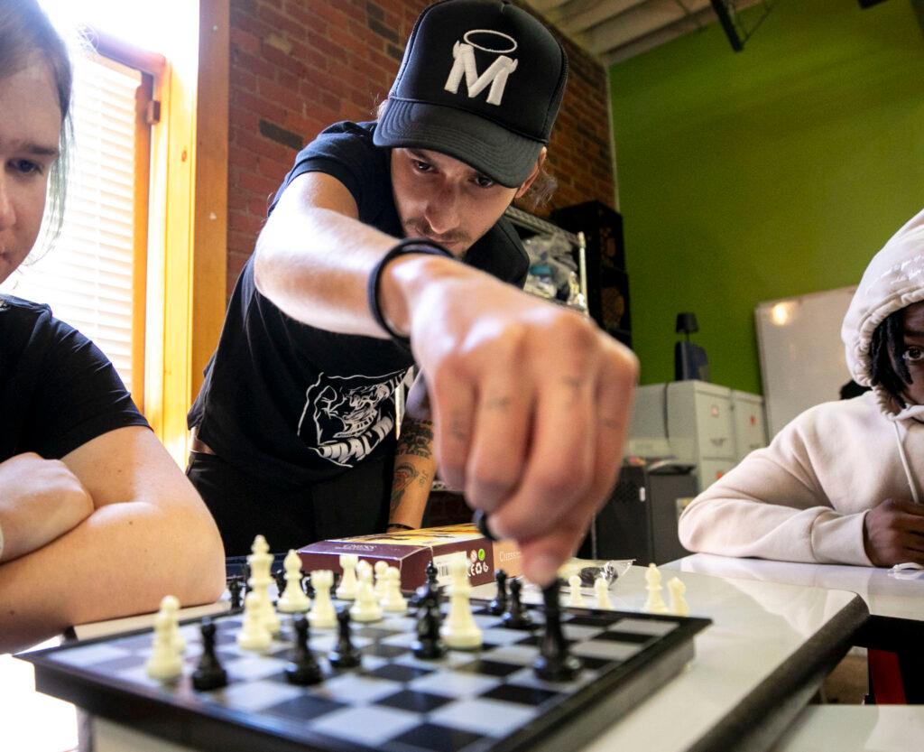 A man in a ball cap reaches forward into the frame, to a chess board on a table. A kid with green-ish hair watches; another kid in a hoodie stares into the lens. A window spills light into the room behind them.