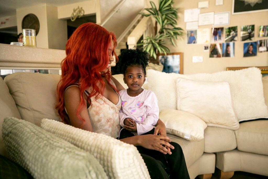 A woman with bright red hair holds a little girl in her lap, in a cream-colored sitting room filled with photos and plants.