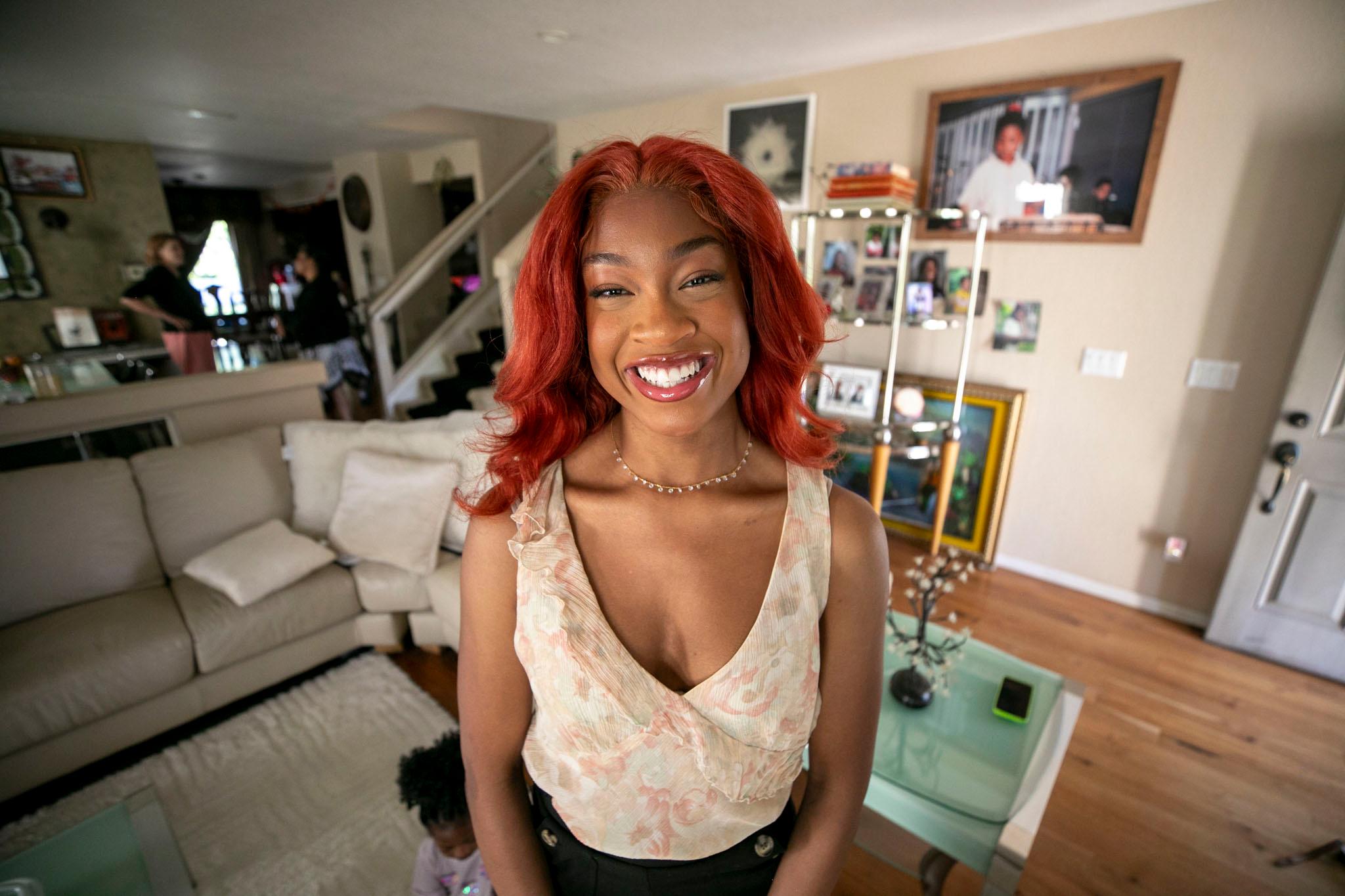 A woman with bright red hair stands in a cream-colored sitting room filled with photos and plants.