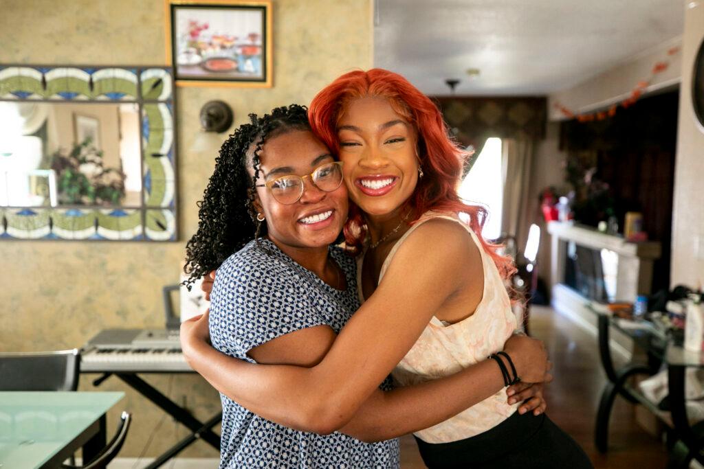 Two women embrace, smiling at the camera, in a brightly lit room.