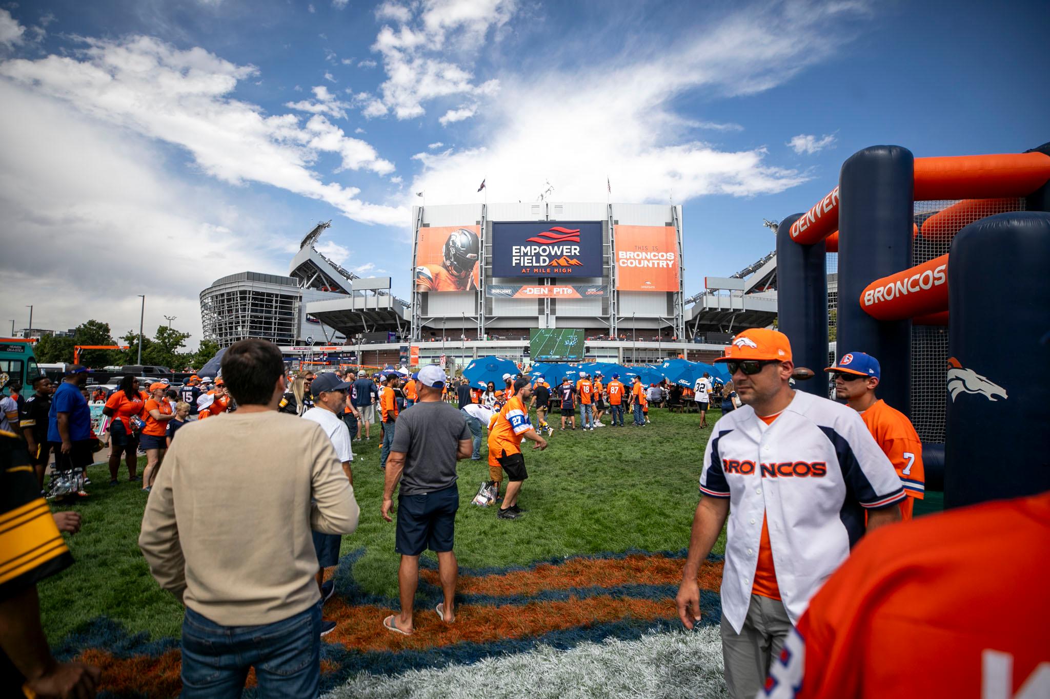 Mile High Stadium, adorned in orange and blue, seen from a lawn outside. People, many in orange jerseys, fill the foreground.