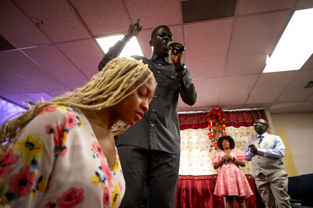 A man in a button-up raises a hand as he yells into a microphone. We're looking up at him, and a woman in a white floral dress kneeling beside him.