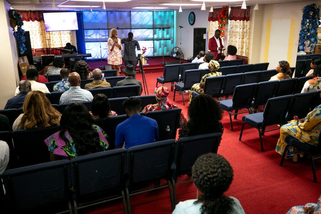 A woman in a white floral dress speaks into a microphone from the front of a red-carpeted room filled with people sitting in rows of chairs.