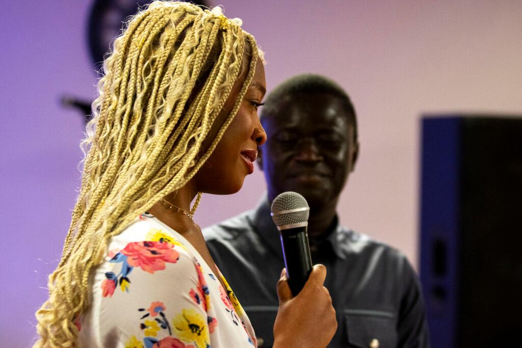 A woman with bleach-blonde braided hair looks emotional as she speaks at a microphone. A man watches from beside her, smiling.