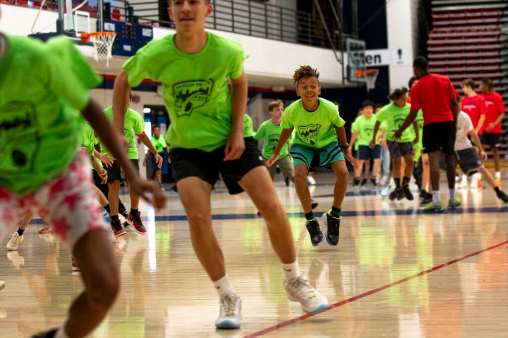 Children in bright green T-shirts skip across the wooden floor of a gymnasium; one in the middle smiles as his feet hover inches above the ground.