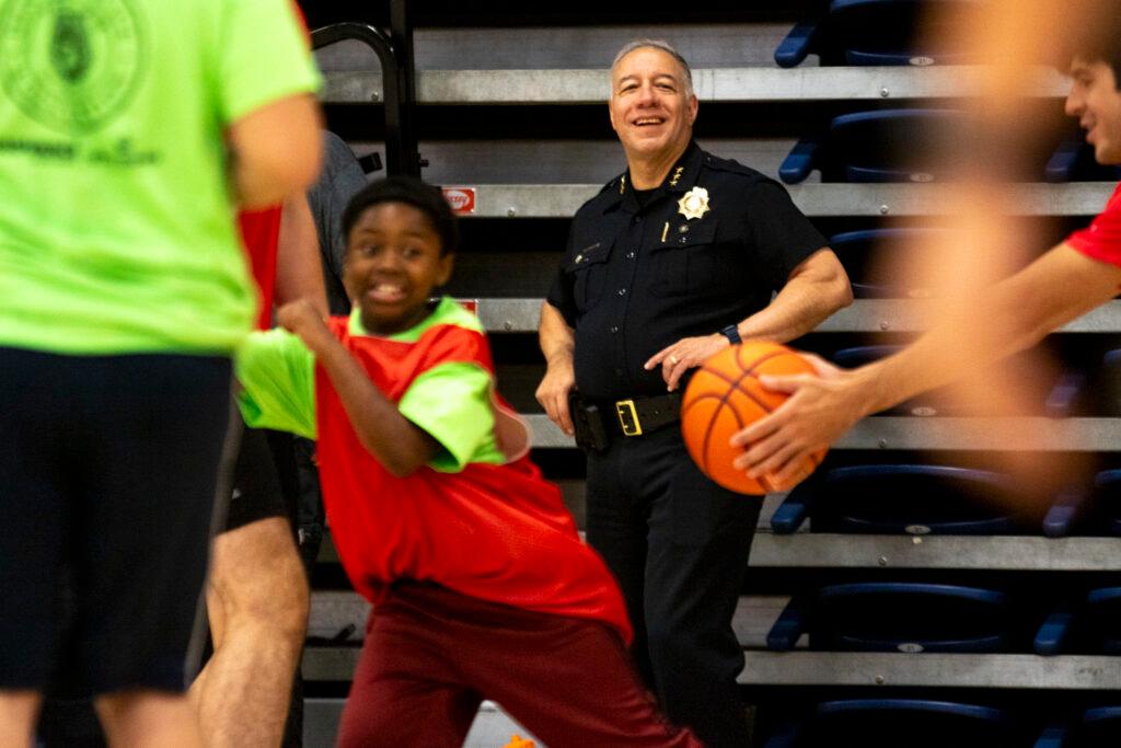 A man in a black police uniform, complete with a shiny gold badge, smiles as children run around a basketball in the foreground.