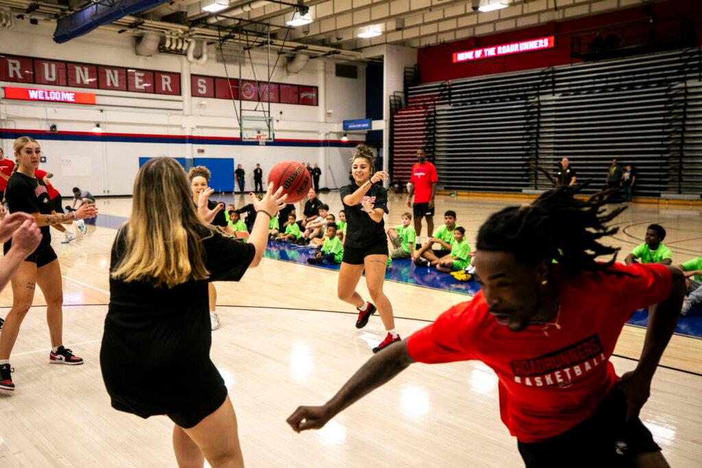 Women in black and men in red sprint around a basketball court; the ball is mid-air in the middle of the frame, as its passed between two players.