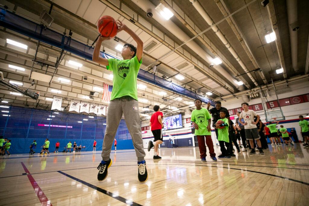 A kid in a lime-green t-shirt floats over a basketball court, mid-jump, as he tosses a basketball over his head. Kids wait in line behind him.