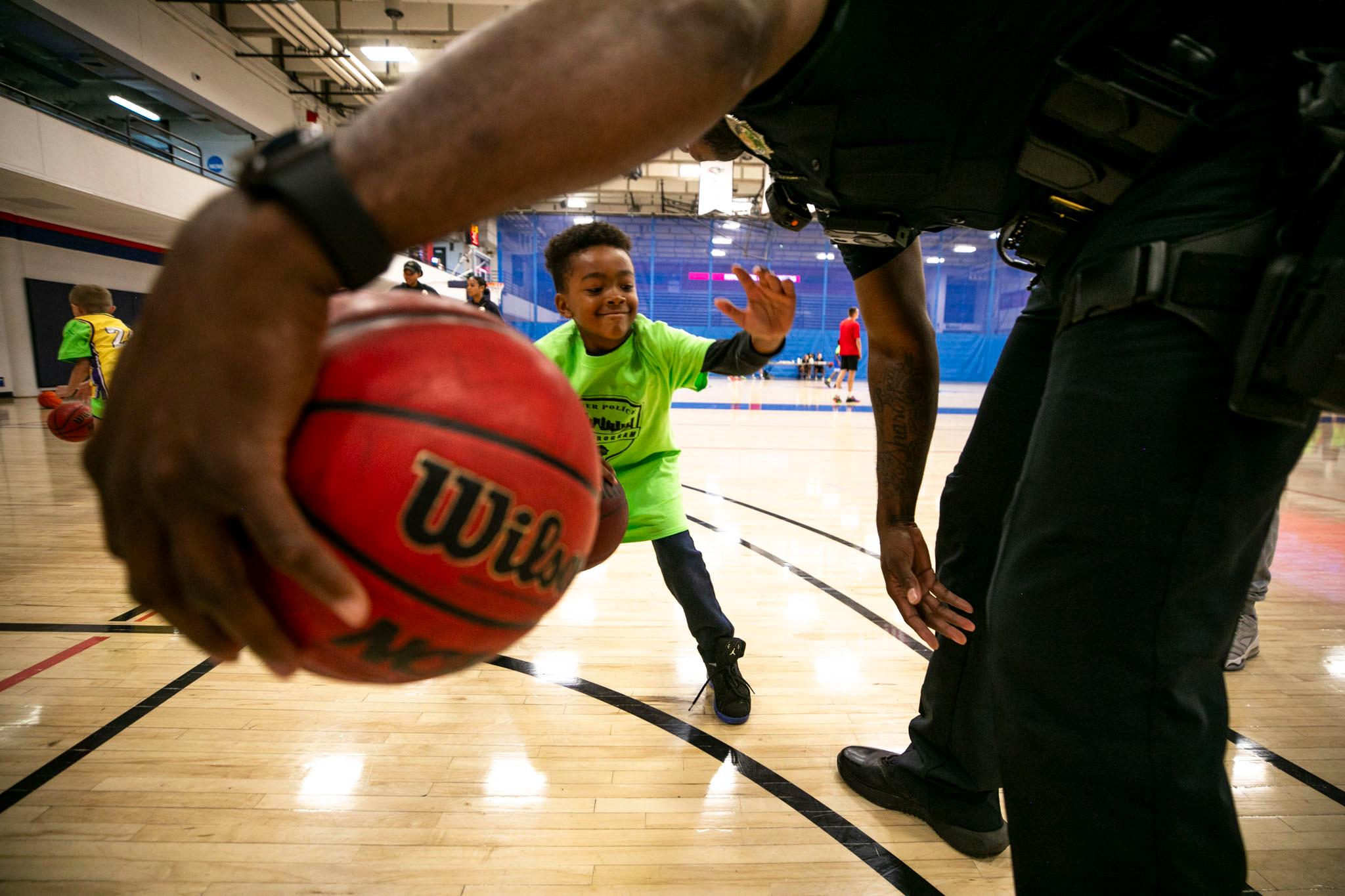 A view under the arm of a man in a black police uniform, which stretches across the top of the frame. His hand holds a basketball as a little kid, seen through the arch of his arm, reaches out to grab it.