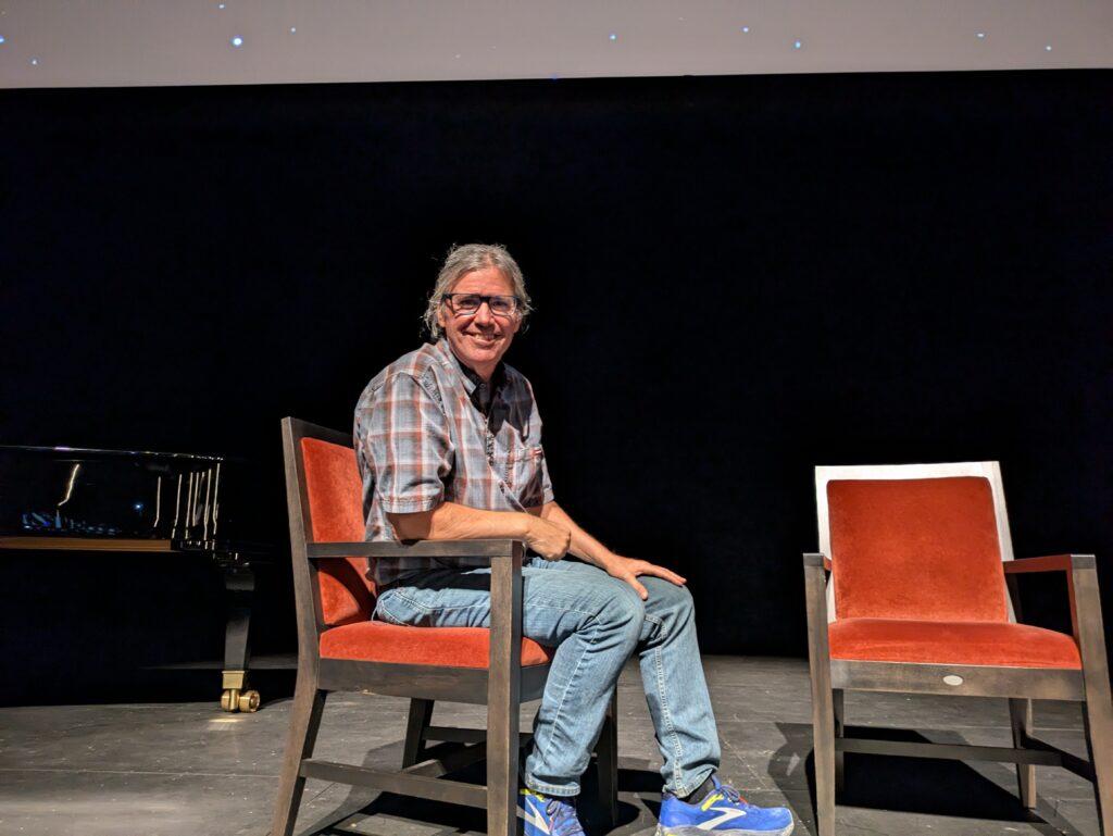 A man in blue-framed glasses with a checkered, short-sleeve shirt poses for a photo while sitting in a red upholstered chair on stage.