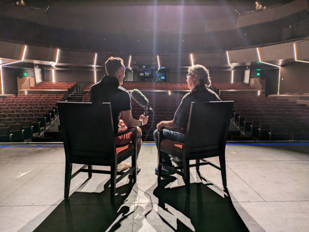 Two men, viewed from behind, sit on stage at an empty theater as stage lighting casts them in silhouette.