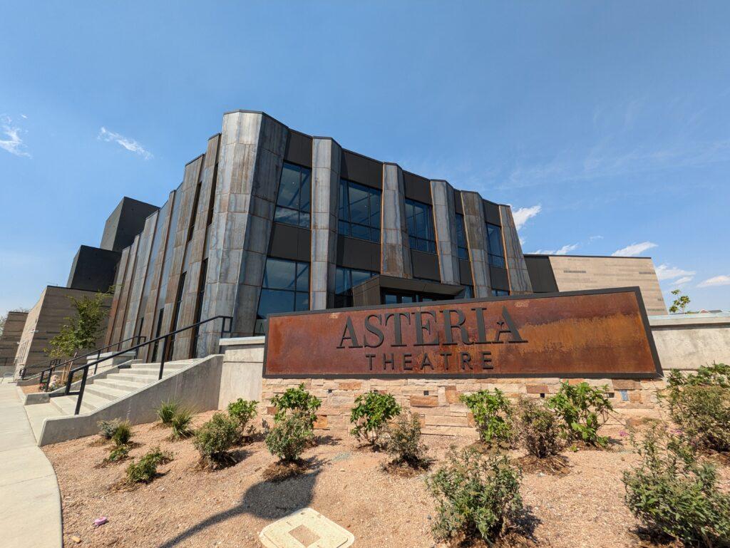 The front façade of Colorado Mesa University's Asteria Theatre is comprised of a multi-story lobby of large glass windows framed by blued steel support.