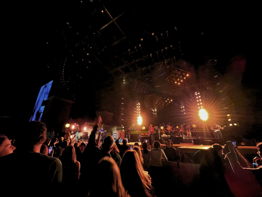 Fans on the floor near the stage watch Cage the Elephant. The sky is dark and the stage light illuminates the tops of people's heads.