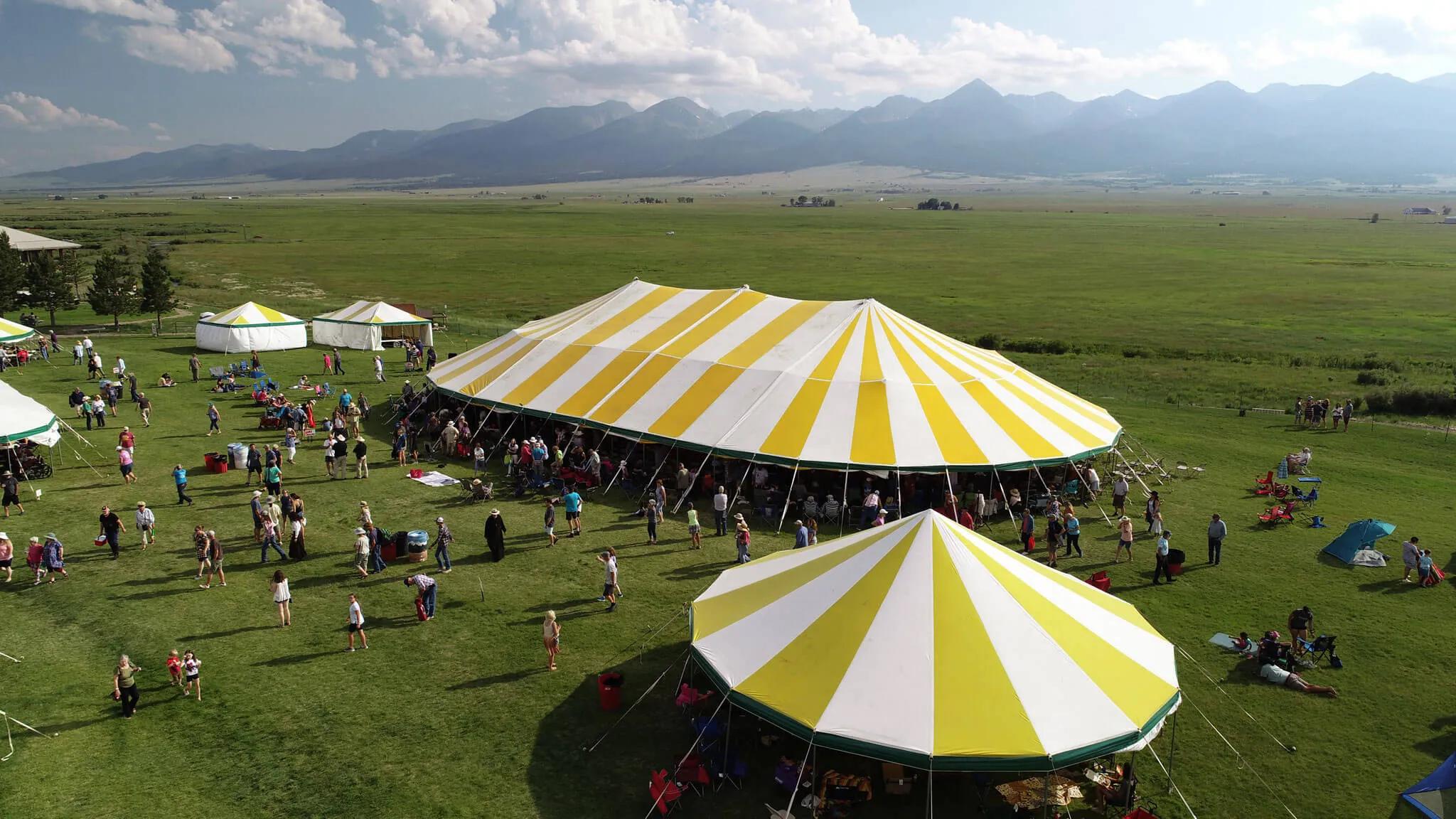 People walk around two large event tents set in a very green meadow with hay fields and mountain views behind them