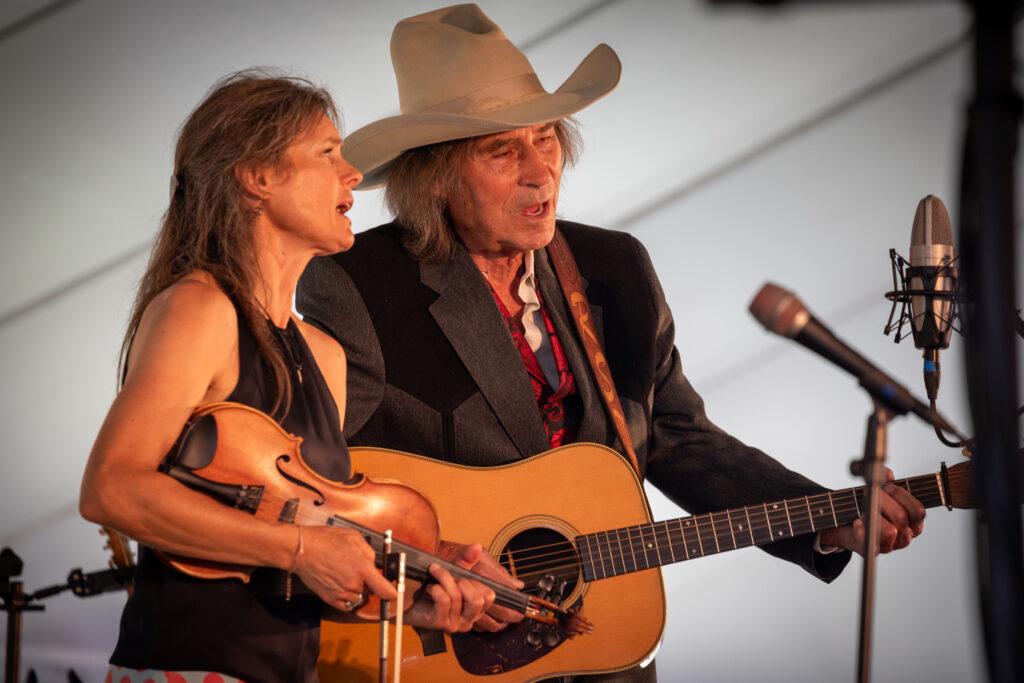 Two people sing into a microphone, The woman is holding a fiddle and the man is playing a guitar.