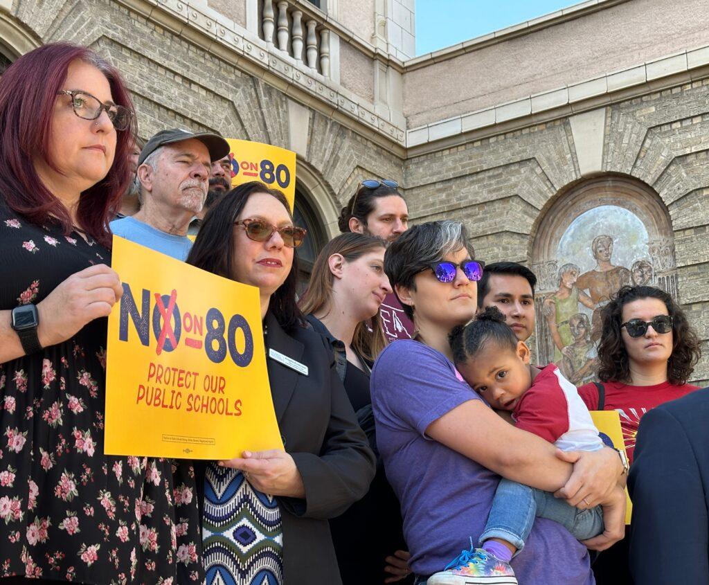 About 10 people stand in front of a white-brick building. One woman is holding her daughter who appears to be about 2-year-old. Two women hold up a sign that reads &quot;No on 80 Protect our Public Schools.&quot;