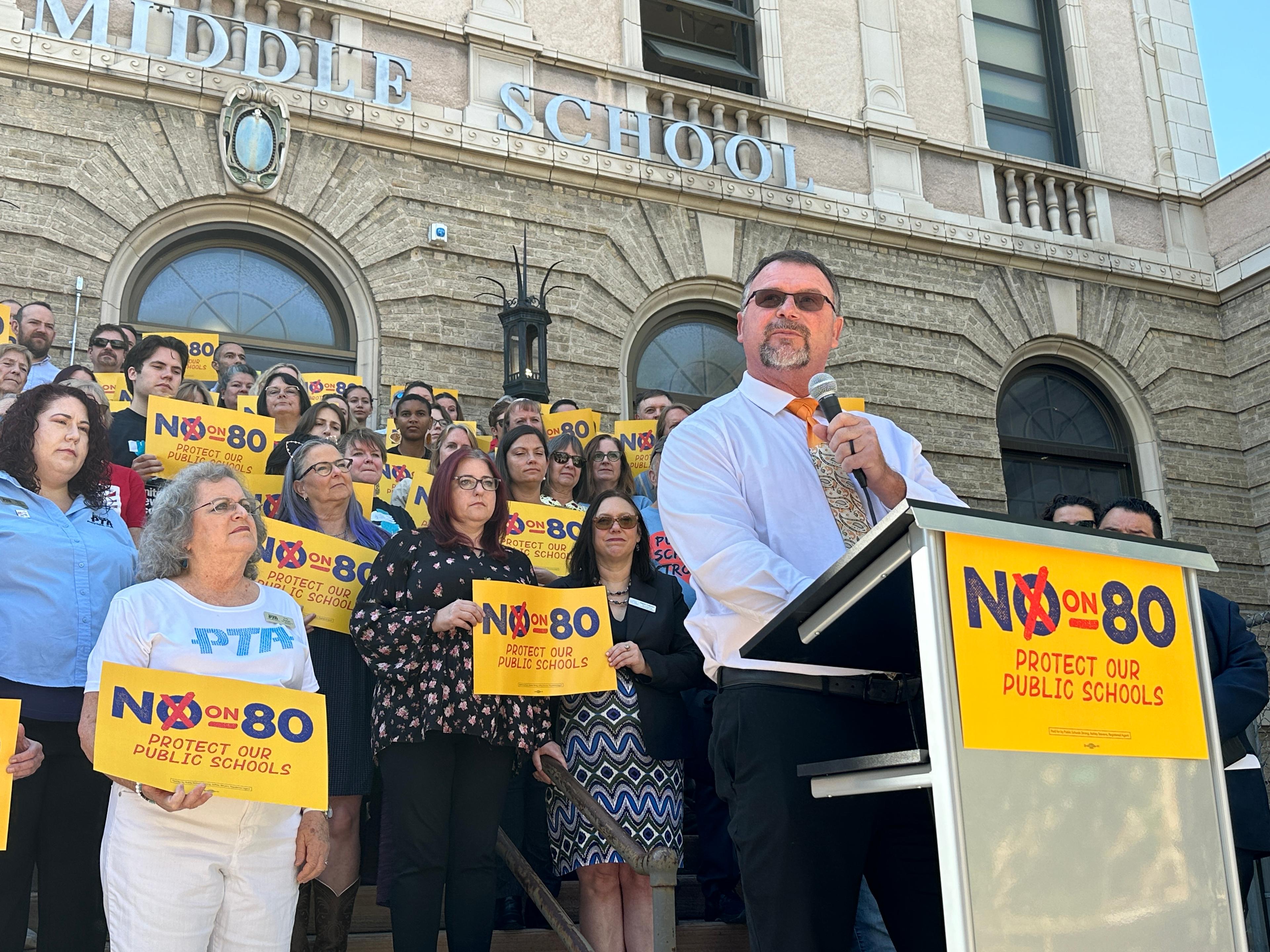 A man stands at a podiun with a microphone in one hand. He is standing in front of a crowd of a few dozen people. The people are holding signs that read "No on 80 Protect Our Public Schools."