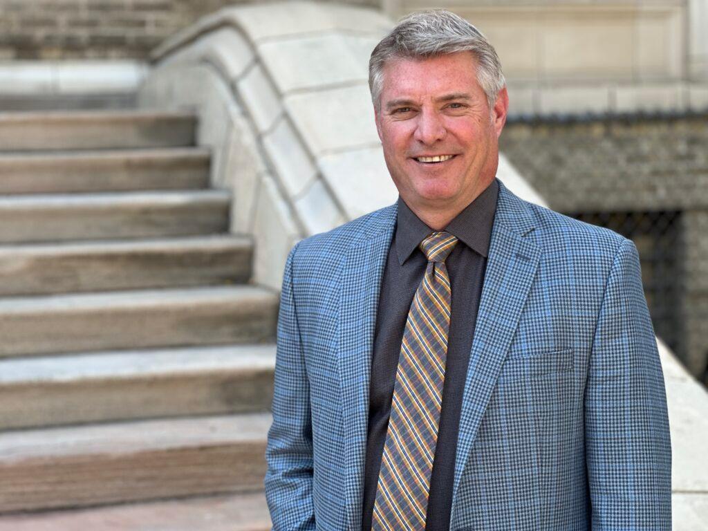 A man with graying hair in a blue dress jacket and a gray and orange plaid tie smile at the camera. In the background are the steps of the historic Morey Middle School