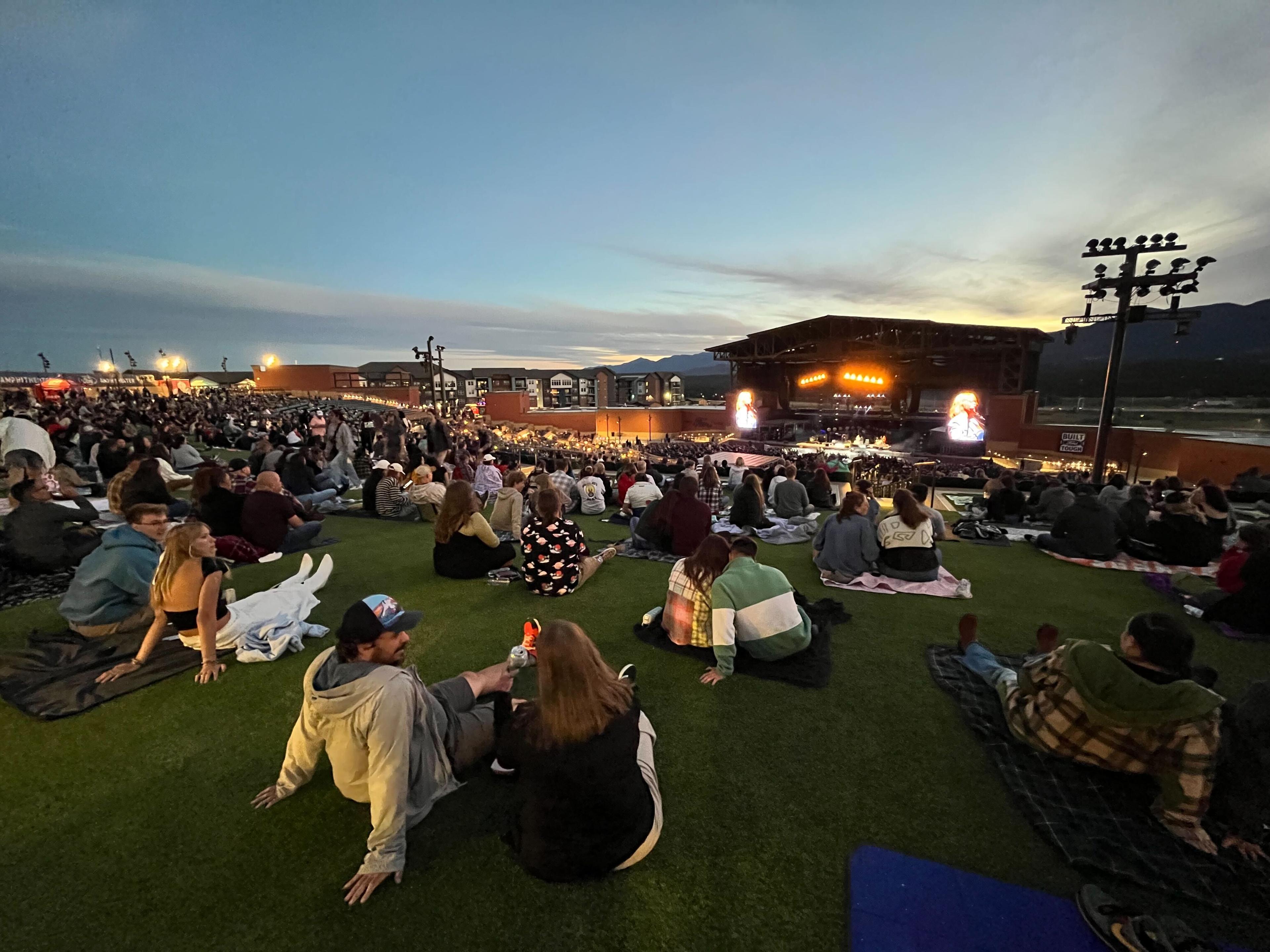 Music lovers gathered on the lawn of Ford Amphitheater for Cage the Elephant. There are many groups of people set up on blankets overlooking the stage with the sun setting in the background.