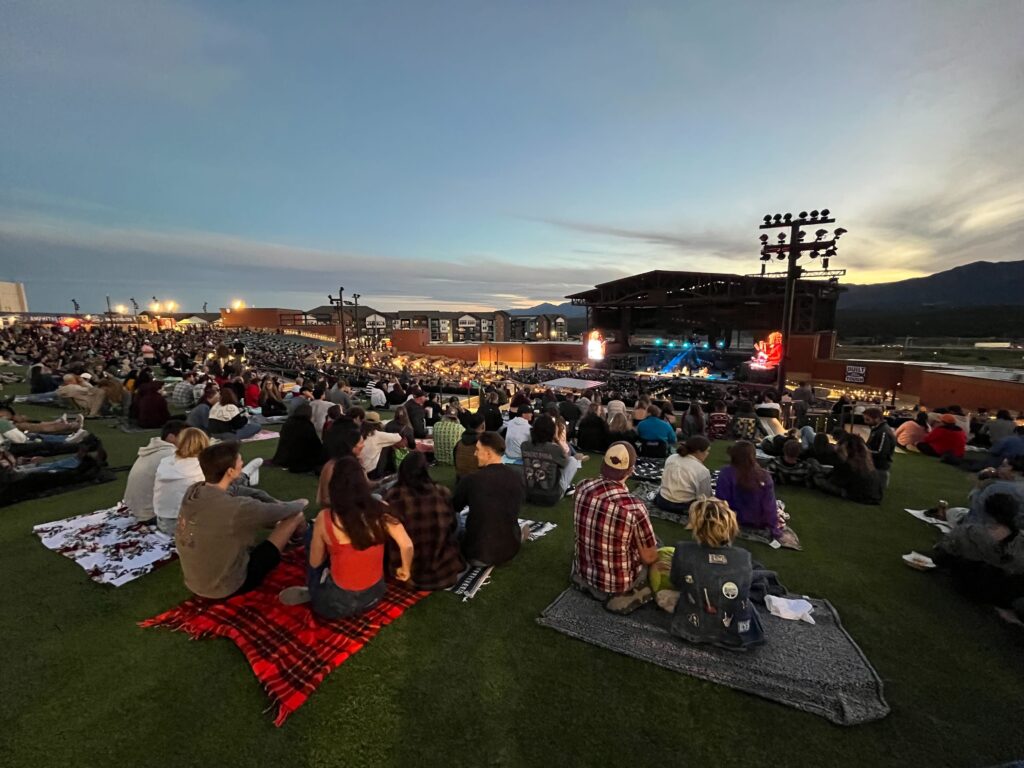 Music lovers gathered on the lawn of Ford Amphitheater for Cage the Elephant. There are many groups of people set up on blankets overlooking the stage with the sun setting in the background.