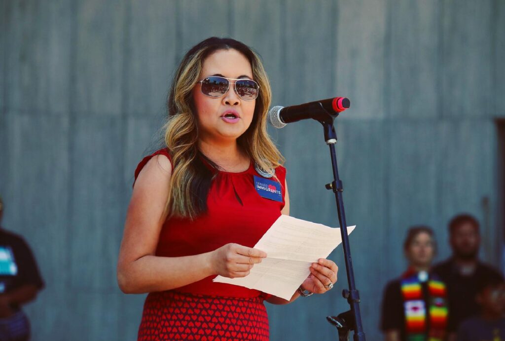 woman in red dress and sunglasses standing at podium giving a speech with notes in her hands.