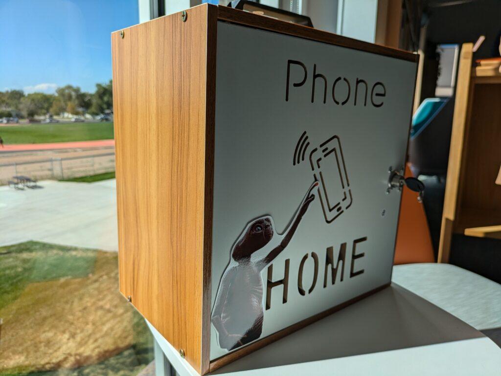 A small wooden box with a lock featuring an aluminum door that has been etched to read &quot;phone home&quot; and features a sticker of the character E.T. from the popular 1982 movie directed by Steven Spielberg.