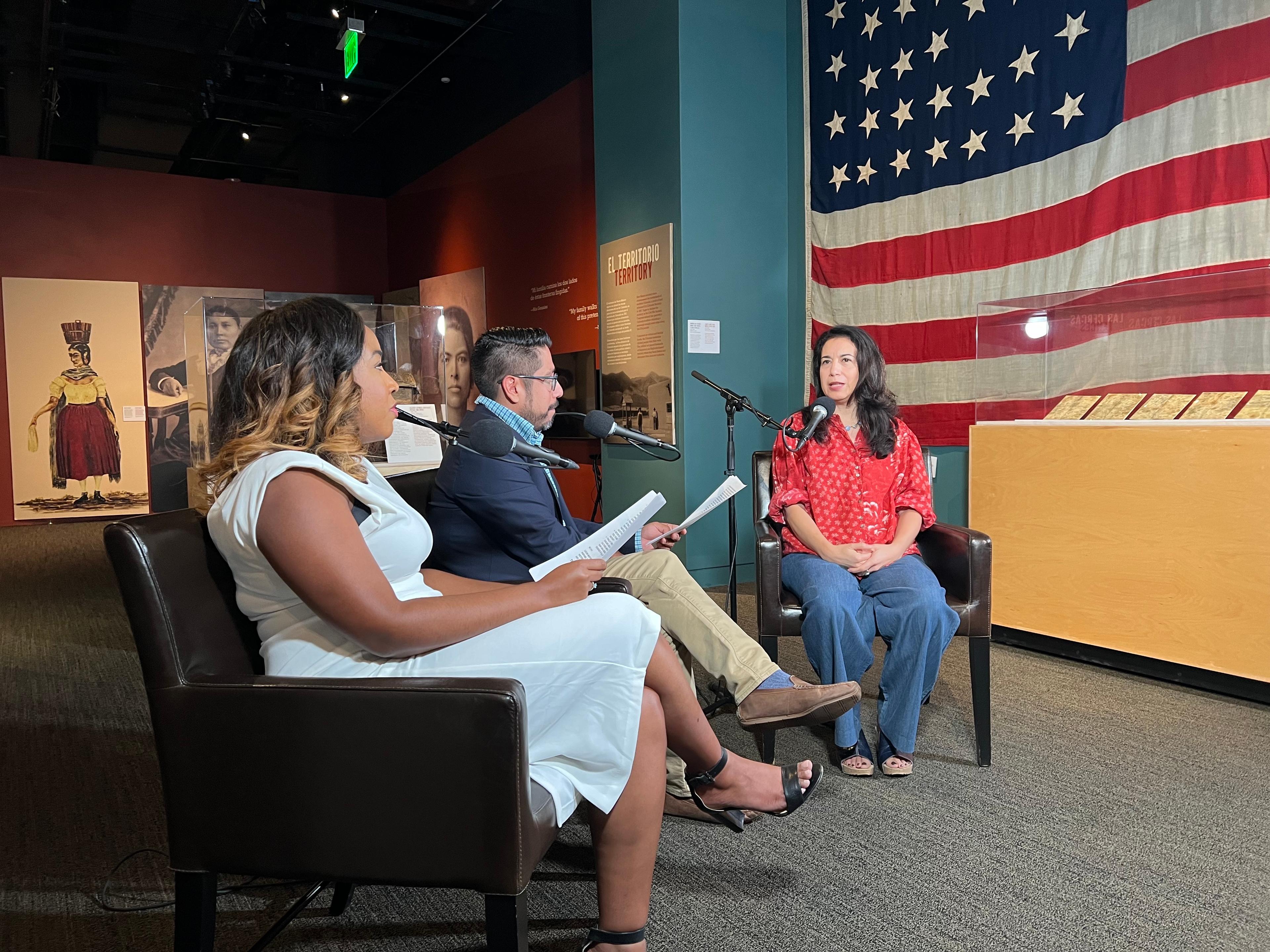 Three people sit in chairs in a museum space with microphones near them. A large American flag is in the background.