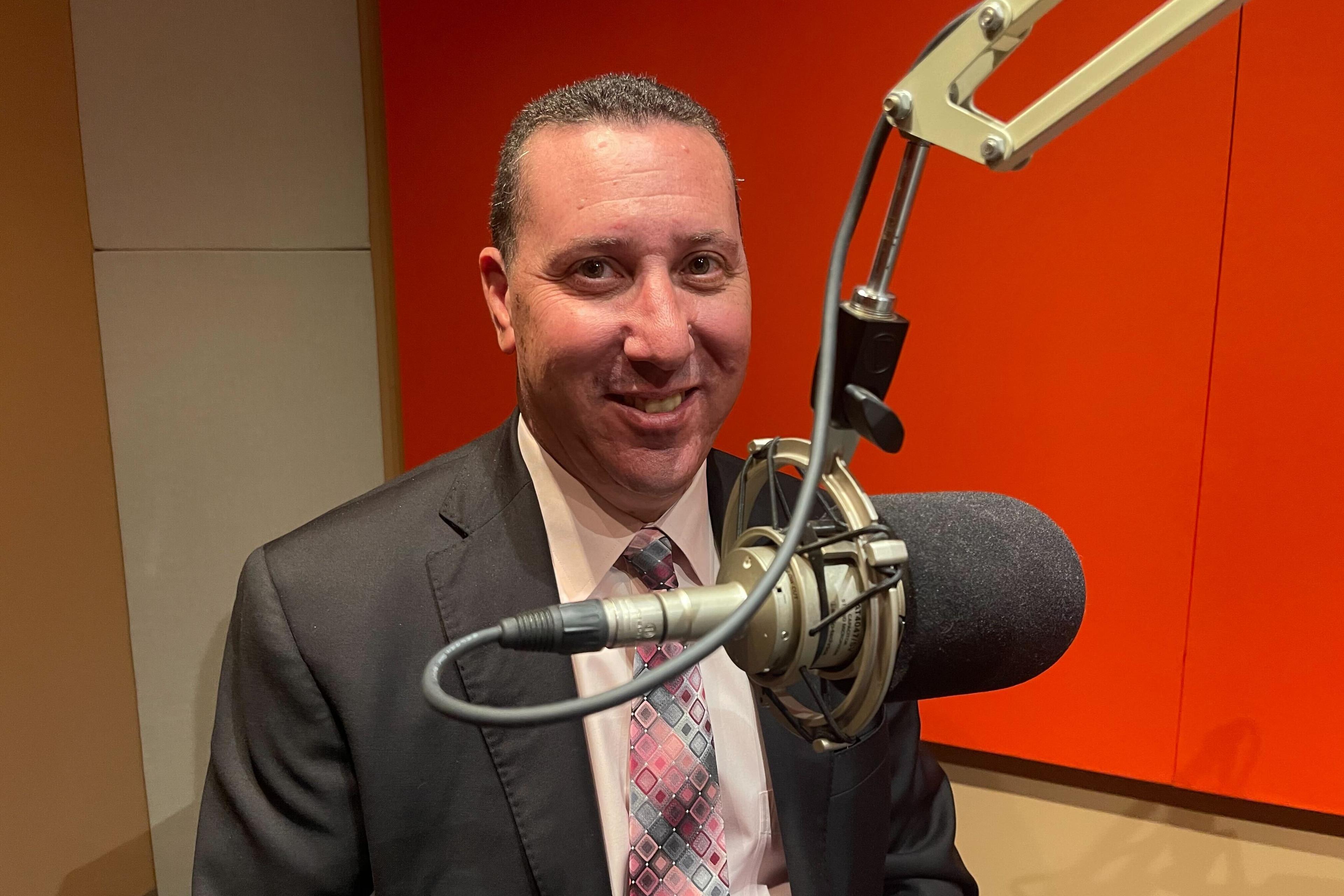 Dan Rubinstein sits in front of a microphone in a sound studio. He is wearing a charcoal suit with a diamond-patterned tie.