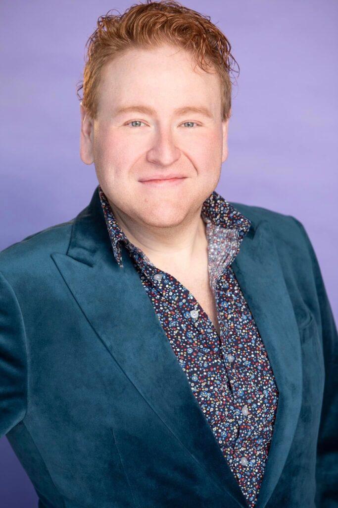 man with red hair and blue velvet blazer over dotted shirt looks smiling into camera