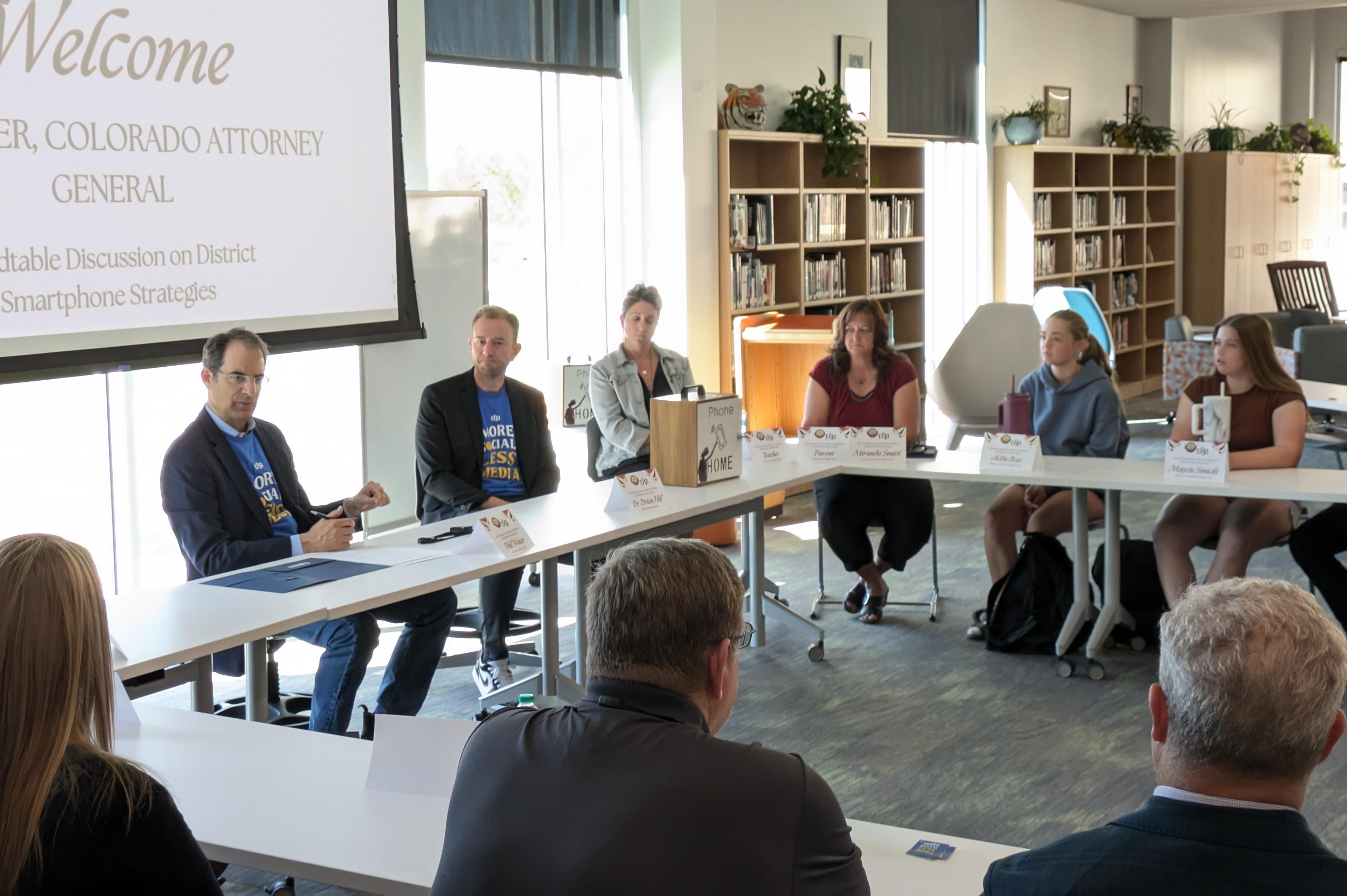 A dozen educators and politicians sit at tables arranged in a circle in a high school library. At the front Attorney General Phil Weiser, wearing a navy blazer and a bright blue t-shirt that says "more social, less media" sits next to Mesa County Valley School District 51 Superintendent Brian Hill, who is wearing a charcoal blazer over a matching "more social, less media" t-shirt.